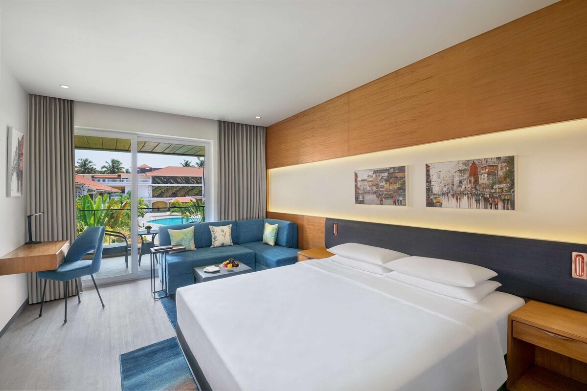 A hotel room with a large bed, a sofa, a desk, and a view of the pool and buildings outside.