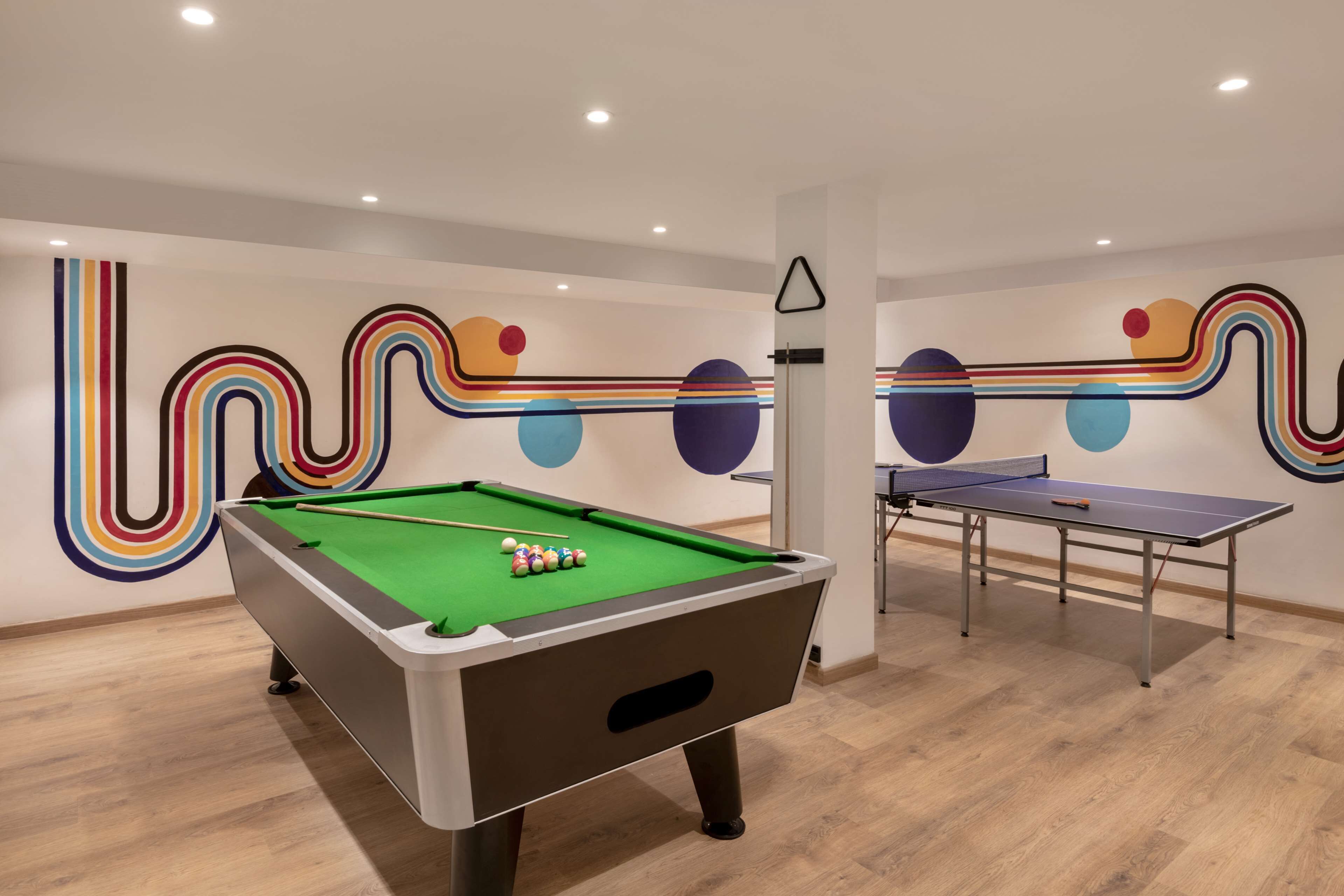 A pool table with balls and a ping pong table in a room with wooden flooring and colorful wall art.