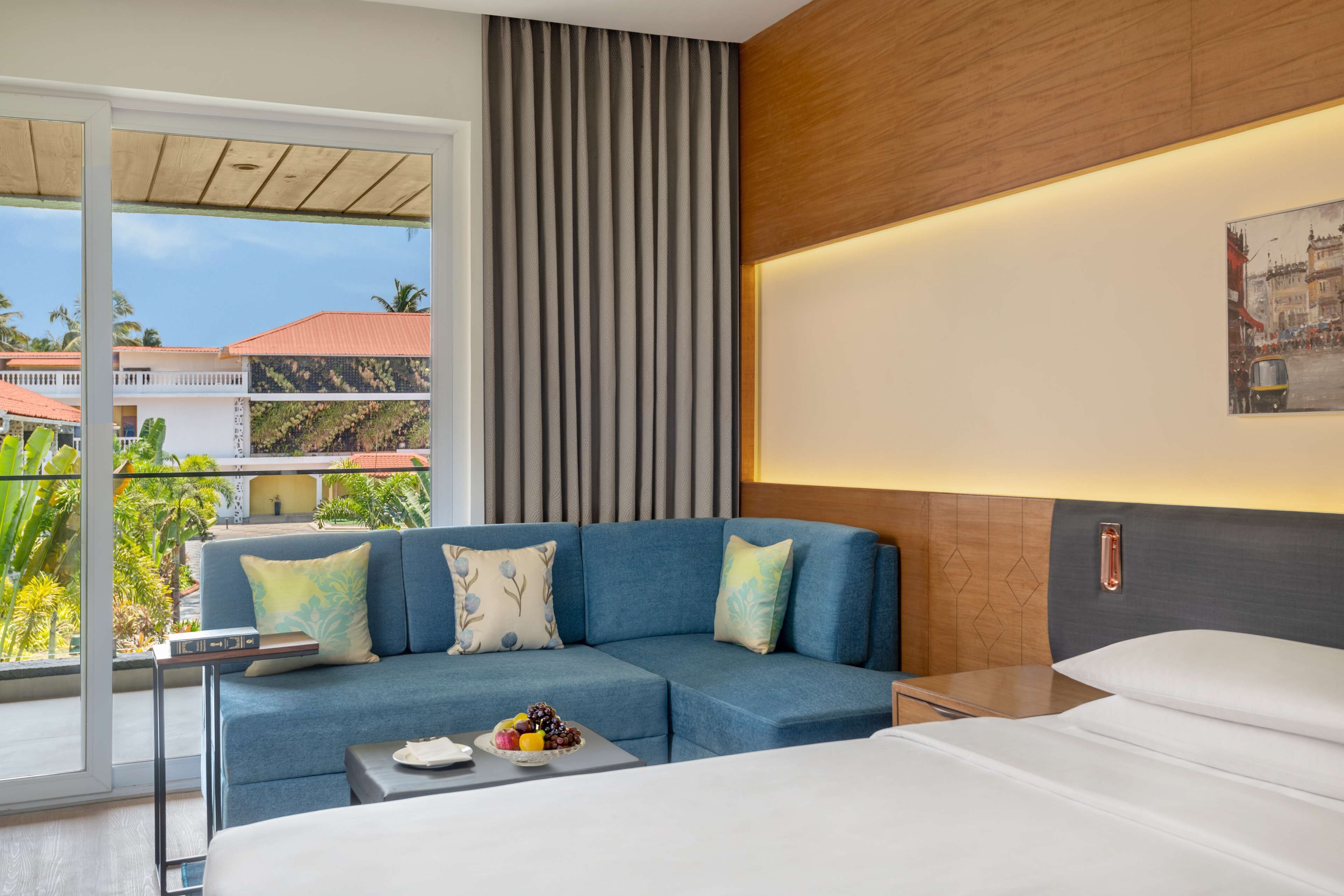 Une chambre d’hôtel moderne avec un canapé bleu, un lit, une petite table avec des fruits et un tableau représentant un paysage urbain accroché au mur.