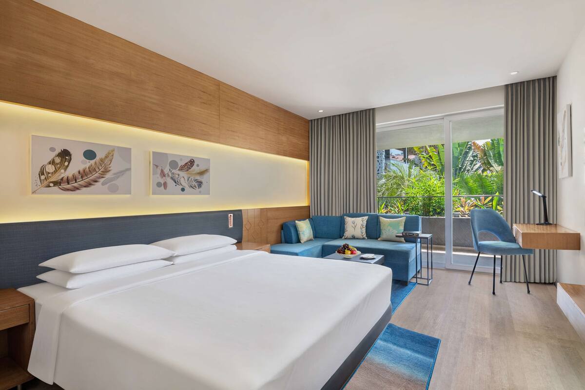 Habitación de hotel moderna con una cama grande, un sofá, un escritorio y un balcón con vistas a zonas verdes.