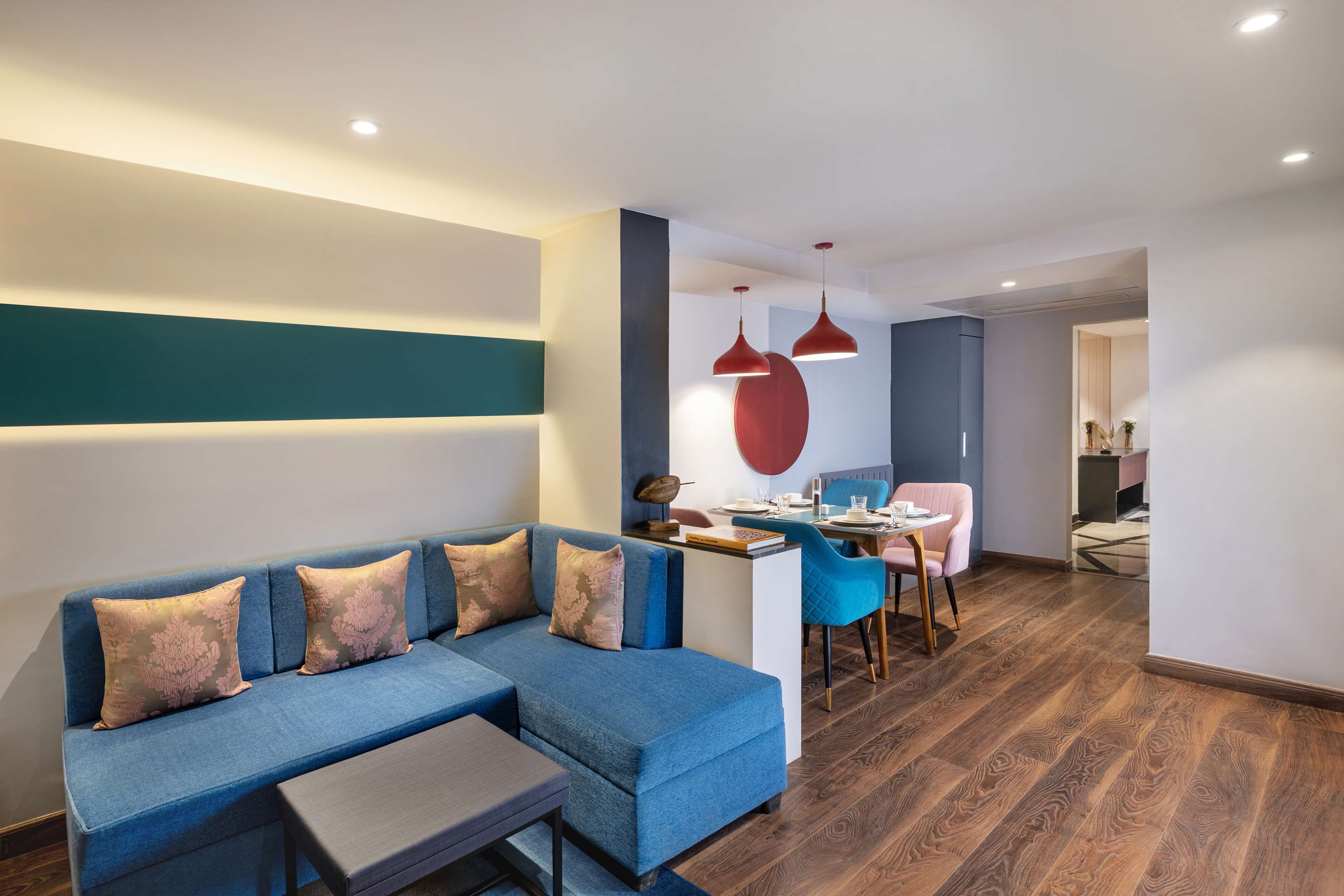A modern living room with a blue sectional sofa, a small table, and a dining area with a pink chair and a blue chair.