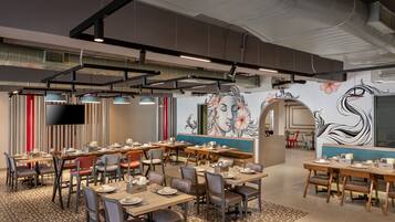 A modern restaurant with multiple dining tables and chairs, a flat-screen TV mounted on the wall, and a decorative mural.