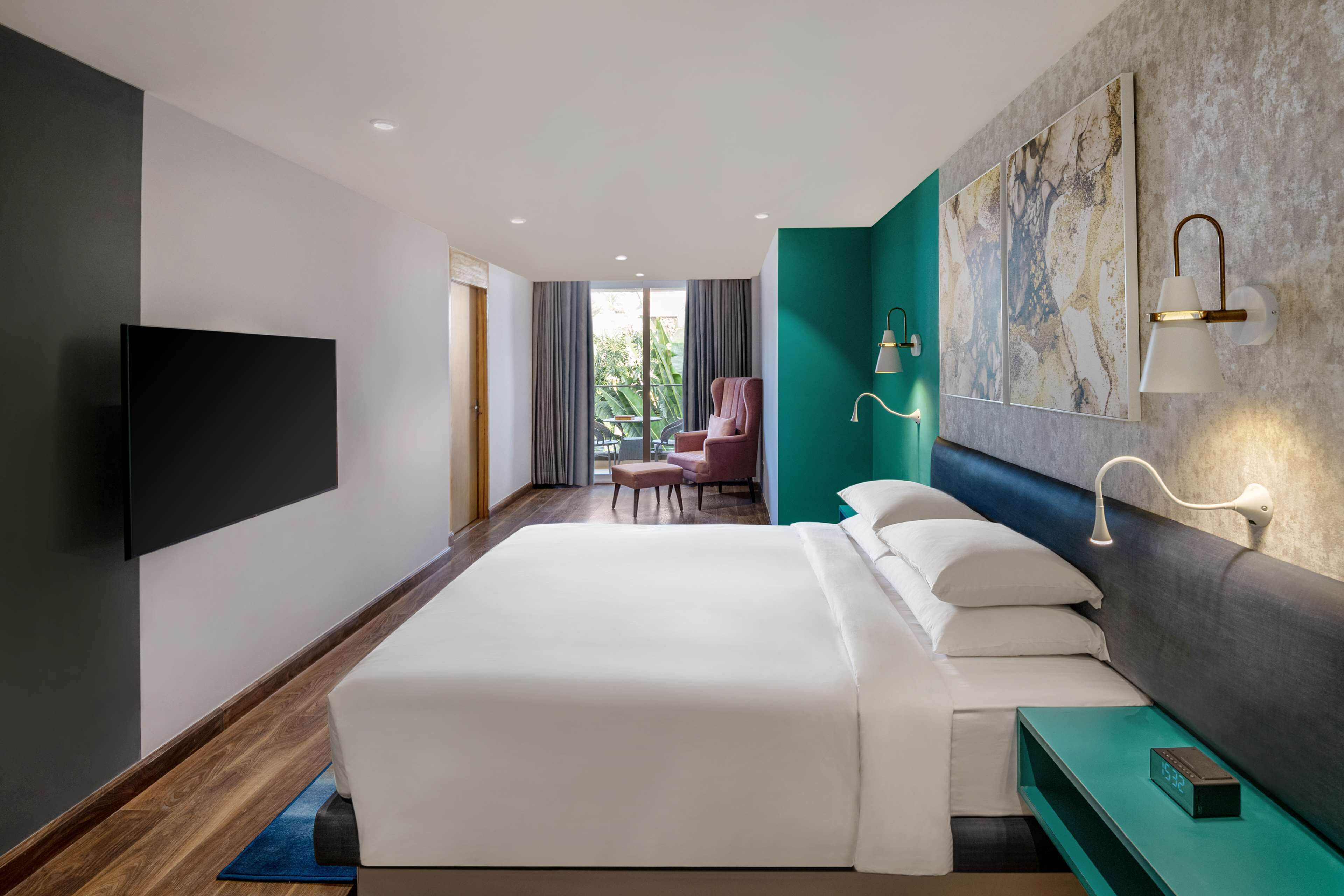 A modern hotel room with a large bed, a flat-screen TV, a teal accent wall, and a view of the outdoors.