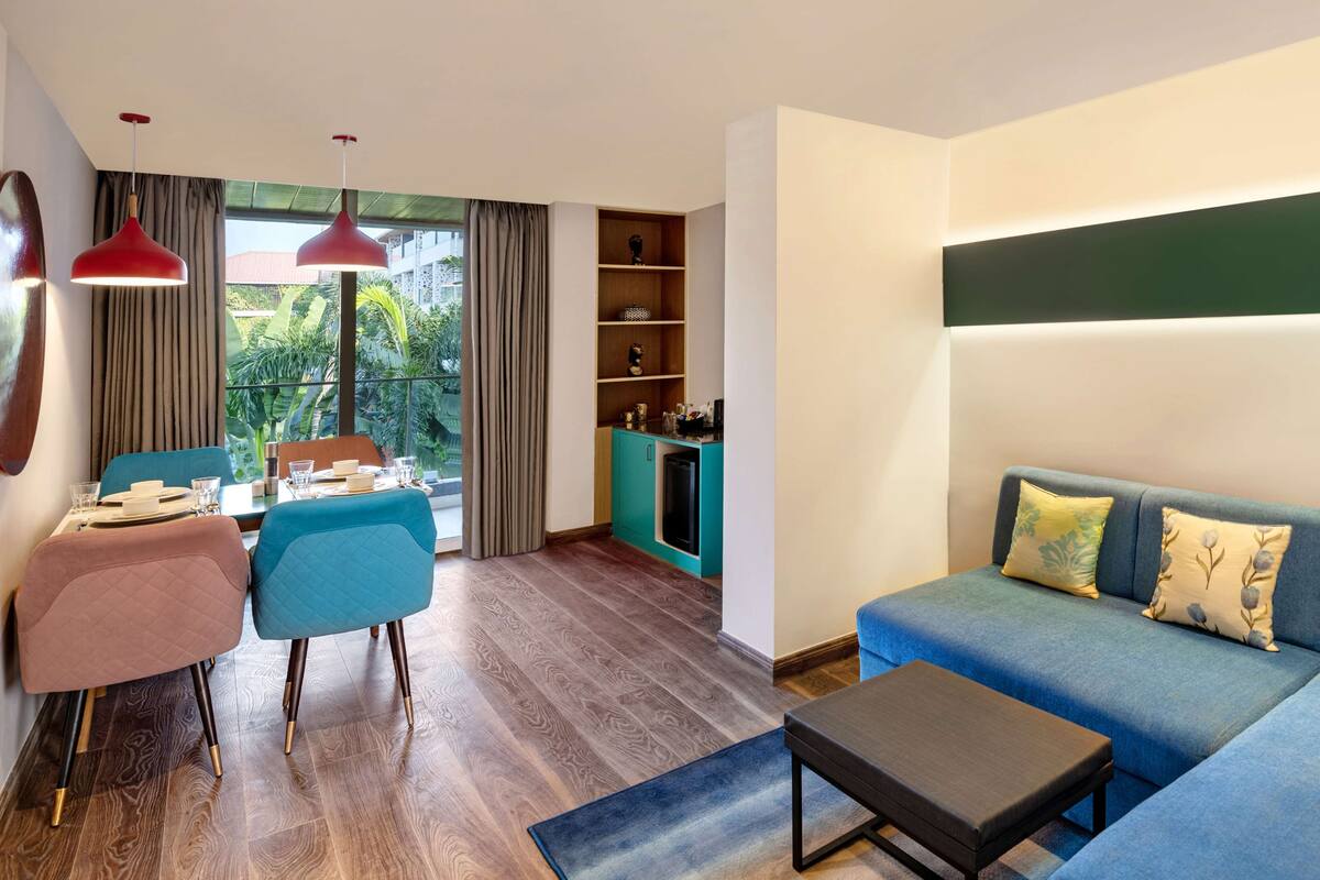 Una sala de estar moderna con sofá, una mesa de centro y un comedor con sillas y mesa. Se observa también una cocina con refrigerador y estanterías.