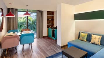 Una sala de estar moderna con sofá, una mesa de centro y un comedor con sillas y mesa. Se observa también una cocina con refrigerador y estanterías.