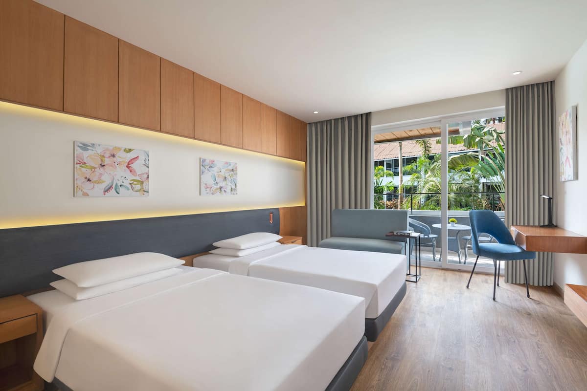 Una habitación de hotel moderna con dos camas, un balcón con mesa y sillas, y vistas a zonas verdes.