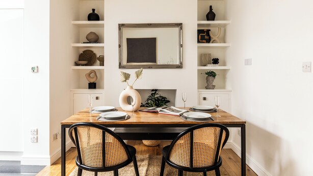 Un coin repas avec une table en bois, deux chaises noires, un miroir au-dessus de la table et des étagères ornées d’objets décoratifs.