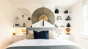 Une chambre à coucher bien rangée, avec un grand lit, un ventilateur fixé au mur et une étagère ornée d’objets décoratifs.