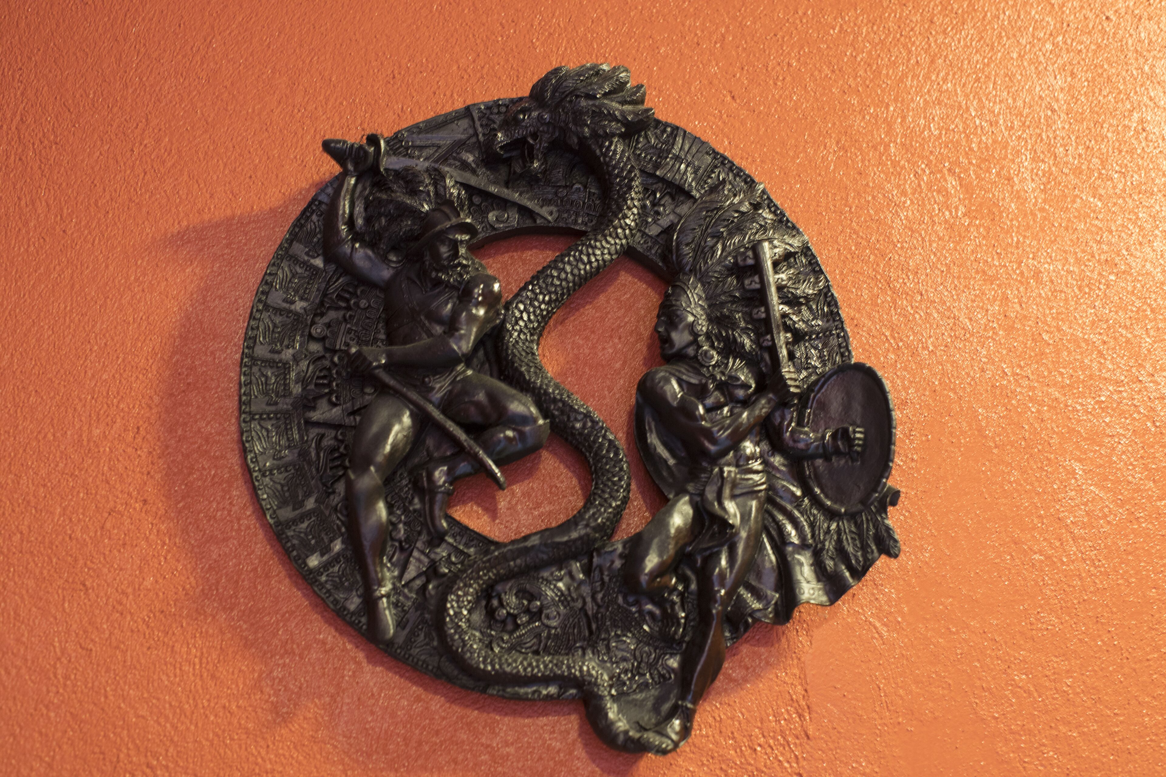 A circular metal sculpture with intricate designs and figures on an orange wall.