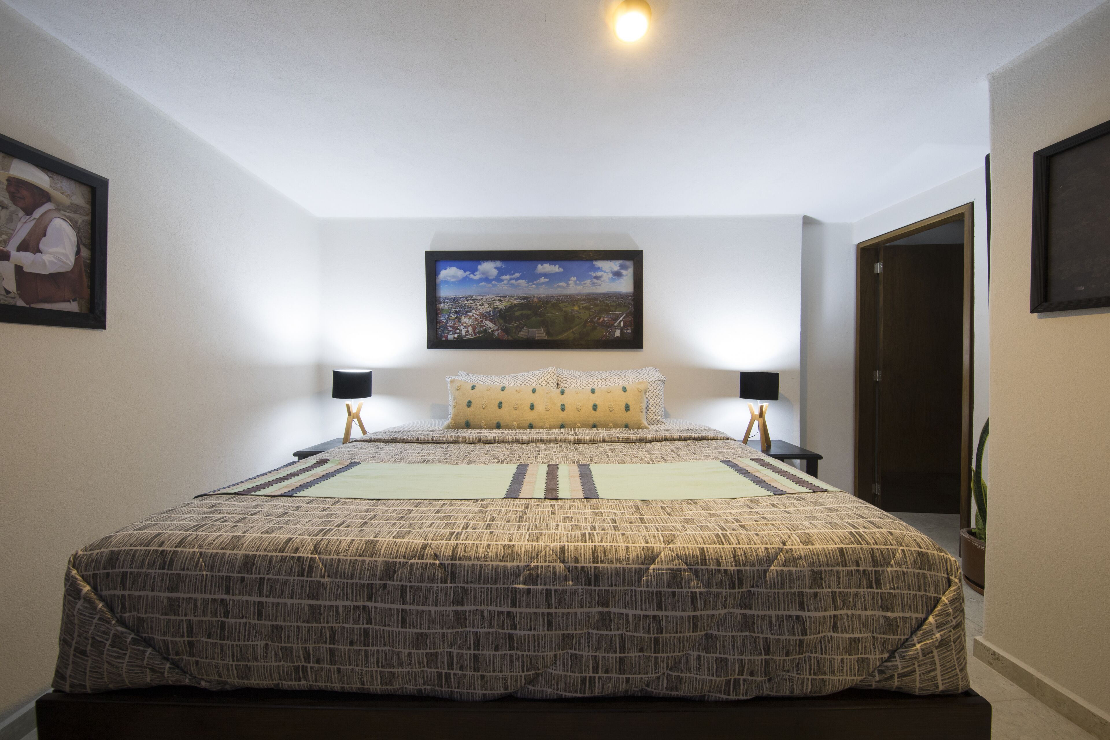 A bedroom with a large bed, two bedside tables, a nightstand, a framed picture on the wall, and a window with a view of a cityscape.