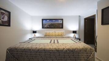 A bedroom with a large bed, two bedside tables, a nightstand, a framed picture on the wall, and a window with a view of a cityscape.