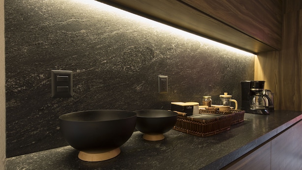 Una cocina moderna con encimeras de granito oscuro, dos cuencos negros, una cafetera y una cesta de pan.