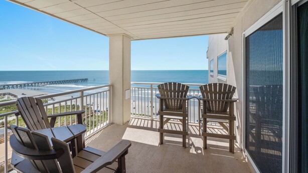 A balcony with two Adirondack chairs and a picnic table overlooking a beach and pier.