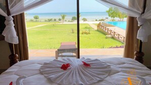 A bed with a white bedspread and a red flower decoration, a view of a pool and a grassy area through a window with sheer curtains. - The Villa of Zanzibar (Kidoti)