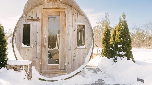Een bolvormige houten hut met een deur en twee ramen, omgeven door sneeuw en bomen.