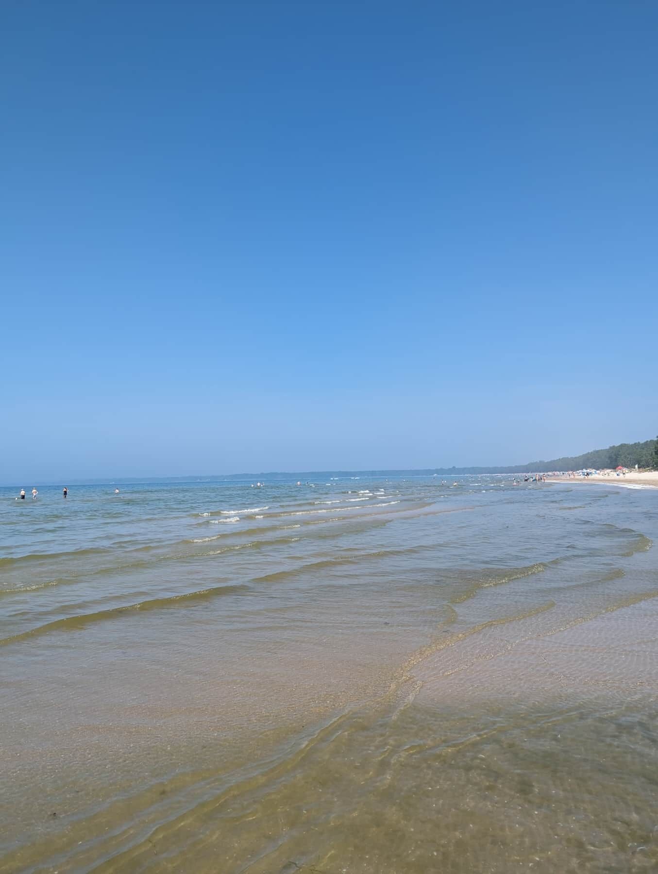 Une plage à l’eau claire, avec un rivage sablonneux et une ligne côtière lointaine où se dressent des bâtiments.