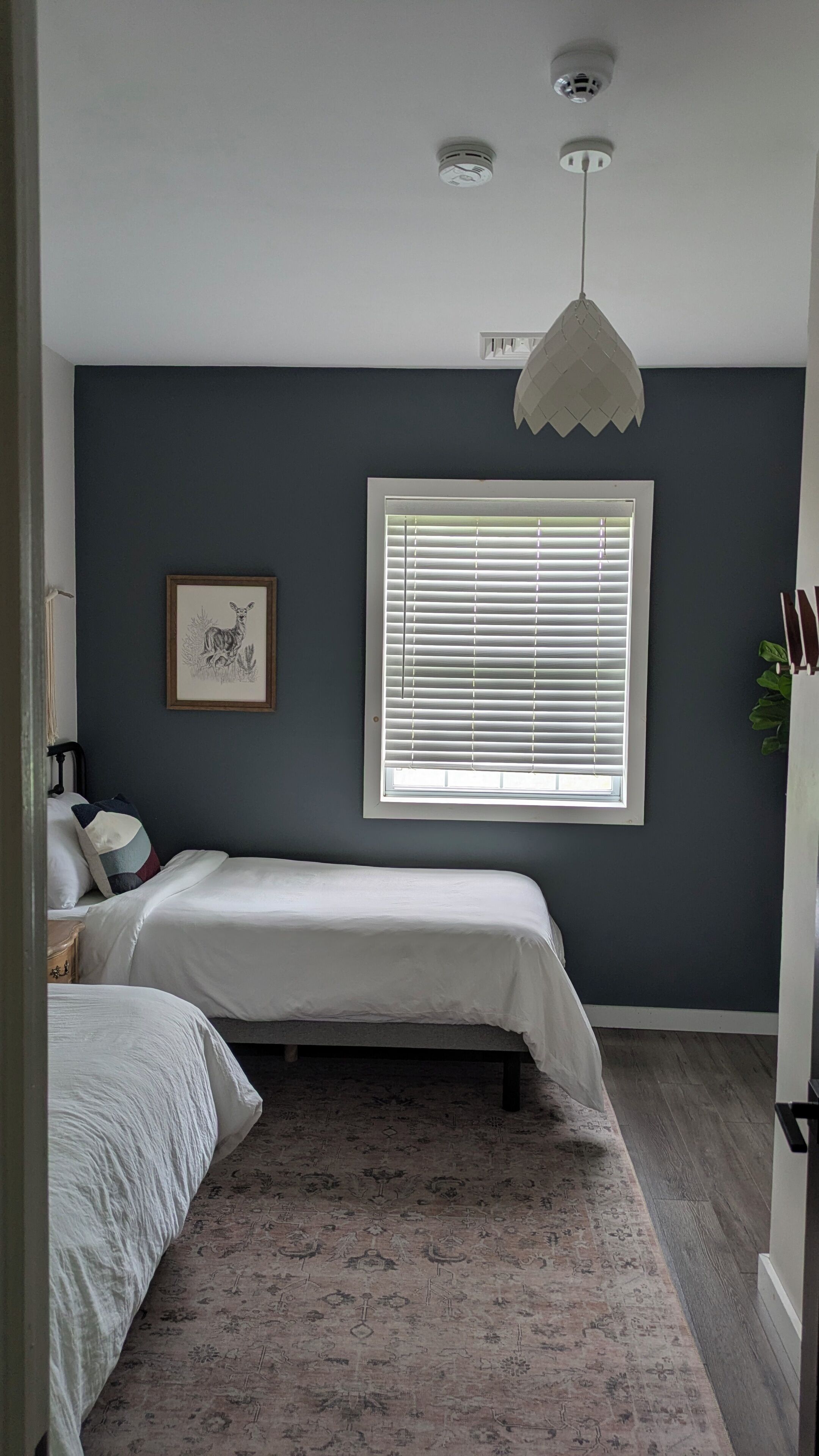 Quarto com cama, janela com persianas, luminária no teto e um quadro pendurado na parede.