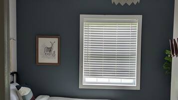 Quarto com cama, janela com persianas, luminária no teto e um quadro pendurado na parede.