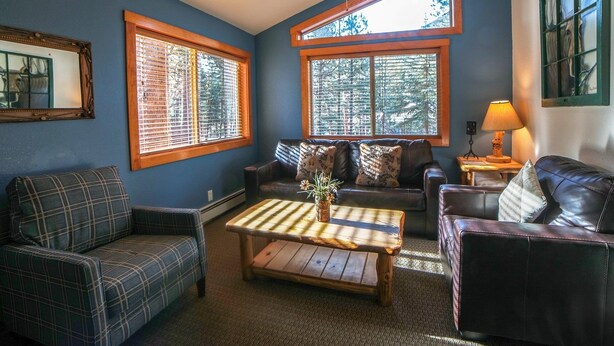 A cozy living room with a blue wall, a wooden coffee table, a sofa, and a leather armchair. There are large windows with blinds, a ceiling fan, and a lamp on a side table. - Timber Creek Chalets: 8 3 Bedroom Chalet (Estes Park)