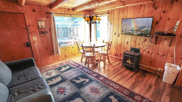 L’intérieur chaleureux d’un chalet, avec un revêtement mural en bois, un coin repas comprenant une table et des chaises, un canapé, une cheminée et un téléviseur fixé au mur.