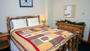 A bed with a quilt featuring animal patterns, a bedside lamp, a mirror, and a wooden dresser with a plant on top. - Timber Creek Chalets: 9 3 Bedroom Chalet (Estes Park)
