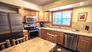 A kitchen with wooden cabinets, stainless steel appliances, a wooden dining table, and a window with blinds. - Timber Creek Chalets: 9 3 Bedroom Chalet (Estes Park)