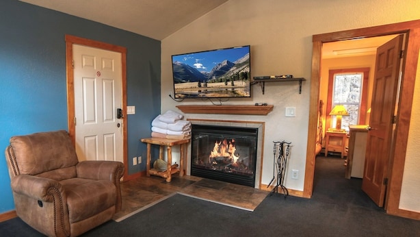 A living room with a fireplace, a television mounted above it, a brown leather chair, and a door leading to another room. - Timber Creek Chalets: 9 3 Bedroom Chalet (Estes Park)