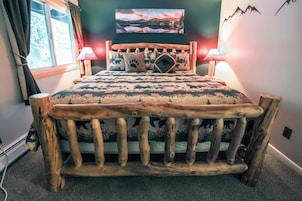 Un letto di legno rustico con biancheria a tema boschivo, due lampade da comodino e un quadro appeso al muro raffigurante un paesaggio montano.
