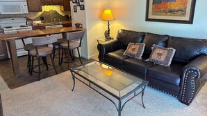 A living room with a black leather sofa, a glass coffee table, and a kitchen area with wooden cabinets and a microwave. - Fawn Valley Inn: 292 1 Bedroom Apts (Estes Park)