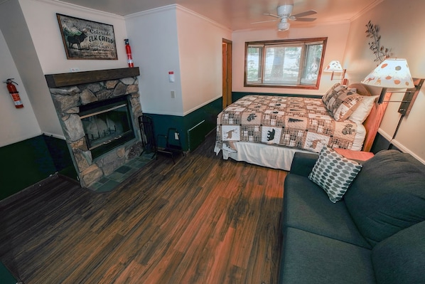 A room with a stone fireplace, a bed with a quilt, a couch, a window with blinds, and a ceiling fan.