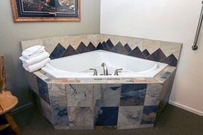Eine Eckbadewanne mit Steinmosaik-Verkleidung, ein gerahmtes Bild darüber und ein Stapel Handtücher daneben.