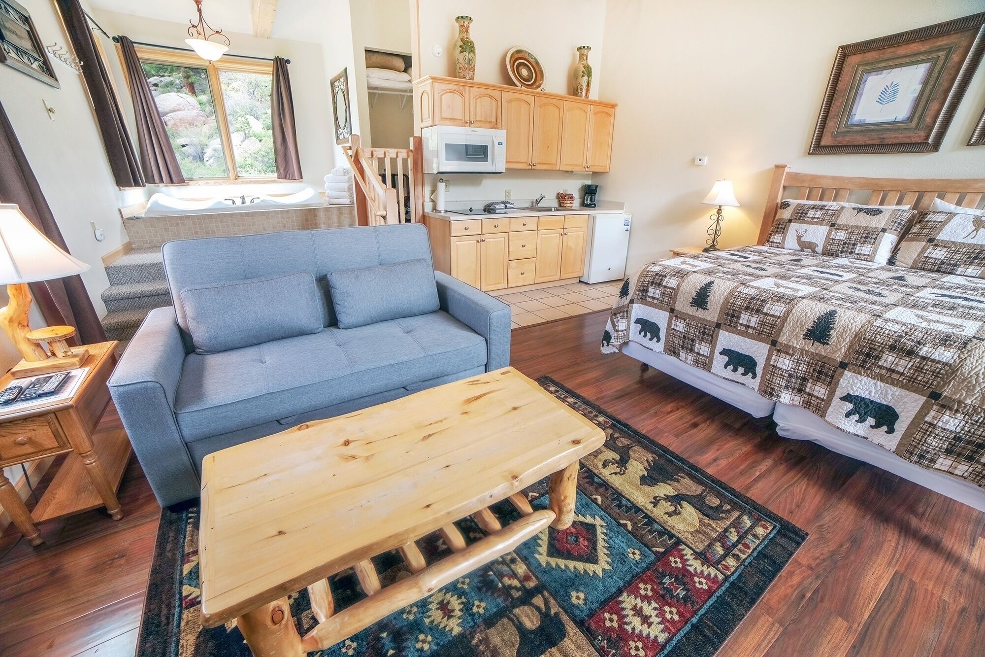 A cozy room with a blue sofa, wooden coffee table, and a bed with a quilted blanket. There is a kitchenette with a microwave and cabinets, and a window with curtains.