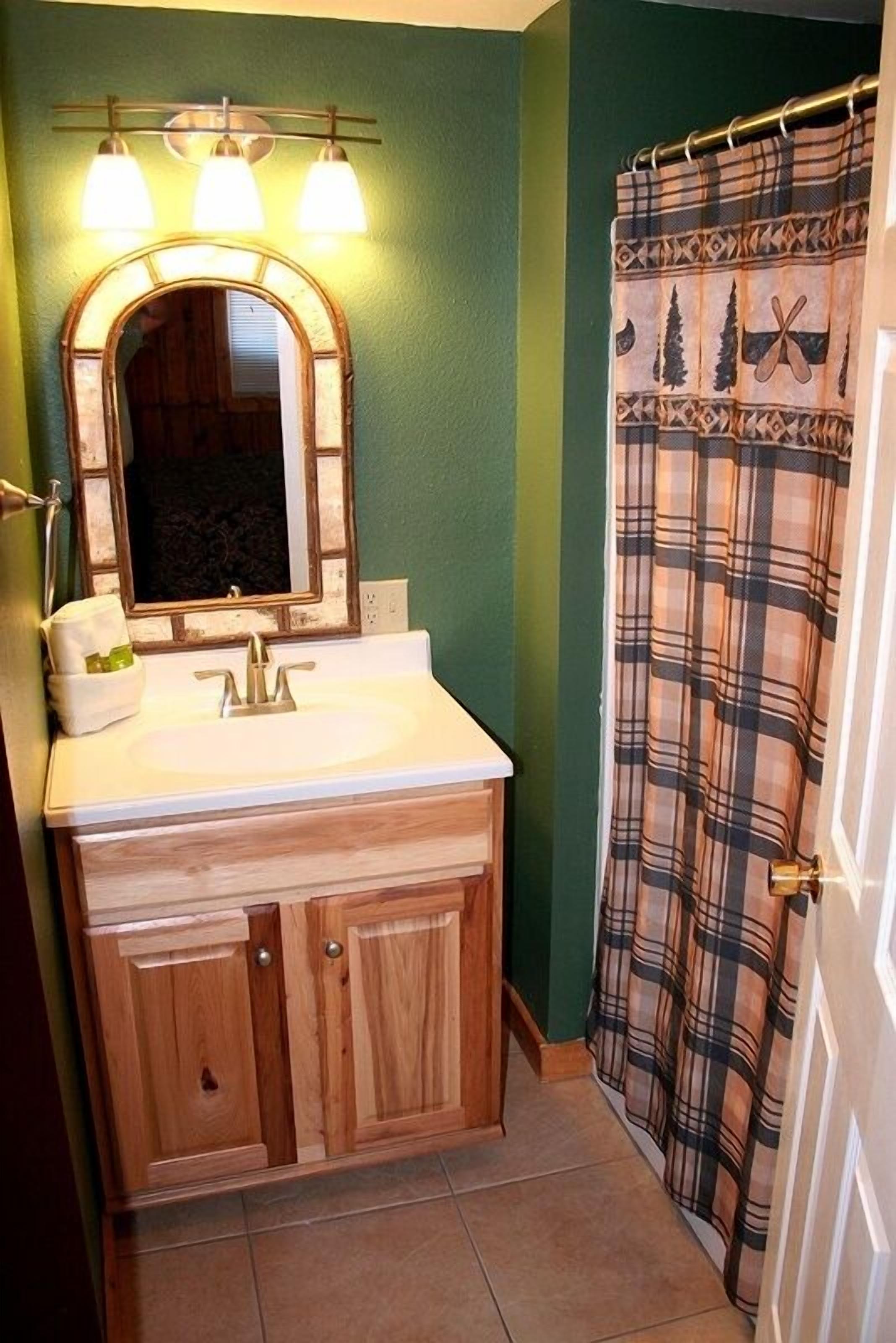 A bathroom with a wooden vanity, a mirror with three lights, and a plaid shower curtain.