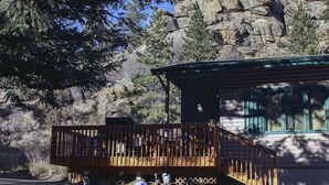 A cabin with a wooden deck, a window with a green frame, and a rocky mountain backdrop. - Sunnyside Knoll: 16 1 Bedroom Cabin (Estes Park)