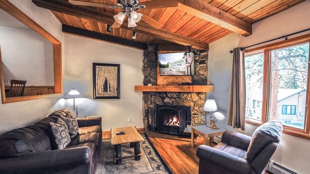 A cozy living room with a fireplace, wooden ceiling beams, and a stone fireplace surround. There is a large window on the right side. - Sunnyside Knoll: 16 1 Bedroom Cabin (Estes Park)