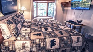 A bedroom with a large bed, a window with curtains, a desk with a chair, a TV on the wall, and a lamp on the nightstand. - Sunnyside Knoll: 16 1 Bedroom Cabin (Estes Park)
