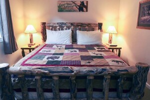 Una cama con colcha estampada con motivos de animales, flanqueada por dos mesitas de noche con lámparas, un cabecero de madera y un cartel enmarcado sobre la cama.