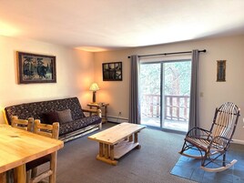 Una sala de estar acogedora con un sofá, una mesa de madera, un sillón mecedor y vistas a través de un ventanal.