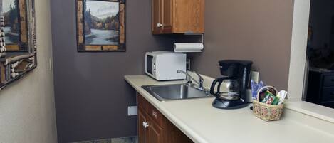 A compact kitchen with a microwave, coffee maker, and a basket of condiments.
