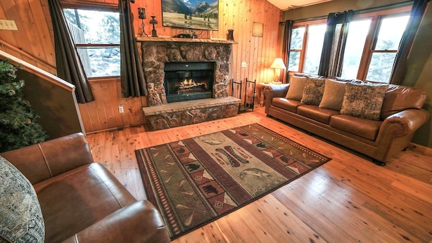 A cozy living room with a stone fireplace, leather sofa, and a large window with curtains. - Lazy R Cottages: 15 2 Bedroom Cabin (Estes Park)