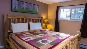 Una cama de madera con colcha, una lámpara de noche, una ventana con cortinas y un cuadro con tema de bosque en la pared.
