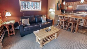 Un espace de vie compact comprenant un canapé, une table basse et des tabourets en bois. Une fenêtre laisse entrer la lumière naturelle, et on aperçoit un coin cuisine.