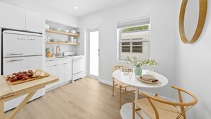 Une cuisine moderne avec des meubles blancs, un réfrigérateur Galanz, une table à manger en bois avec des chaises, et une petite fenêtre donnant sur un espace verdoyant.