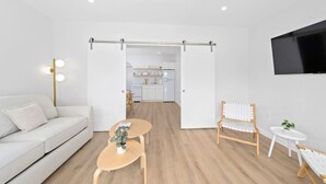 A modern living room with a sofa, a coffee table with a plant, a wooden chair, a small round table, and a TV mounted on the wall.