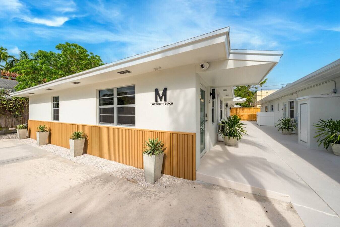 A modern building with a 'M' logo and 'LAKE WORTH BEACH' text, featuring a covered outdoor area and potted plants.