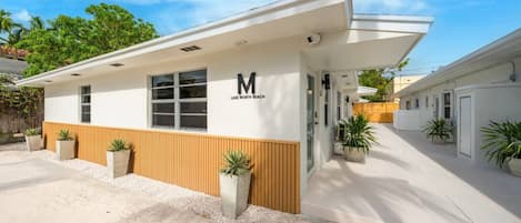 A modern building with a 'M' logo and 'LAKE WORTH BEACH' text, featuring a covered outdoor area and potted plants.