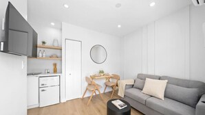 Un espacio habitable moderno y minimalista con un sofá gris, una mesa de comedor con sillas, una pequeña cocina y un espejo redondo en la pared.