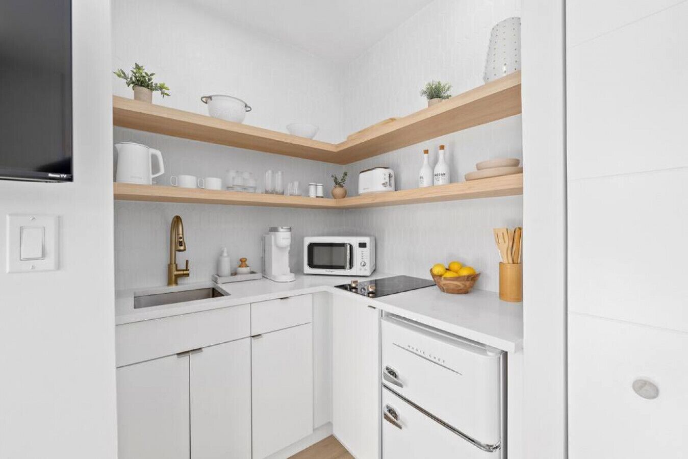 Een kleine keuken met witte kastjes, een magnetron, een koffiezetapparaat en een spoelbak. Daarboven hangen houten planken met verschillende keukenspullen.