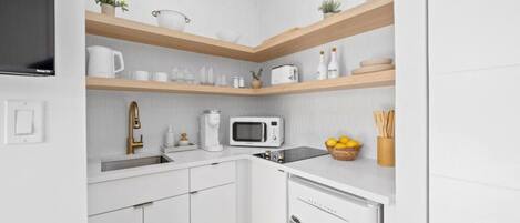 Een kleine keuken met witte kastjes, een magnetron, een koffiezetapparaat en een spoelbak. Daarboven hangen houten planken met verschillende keukenspullen.