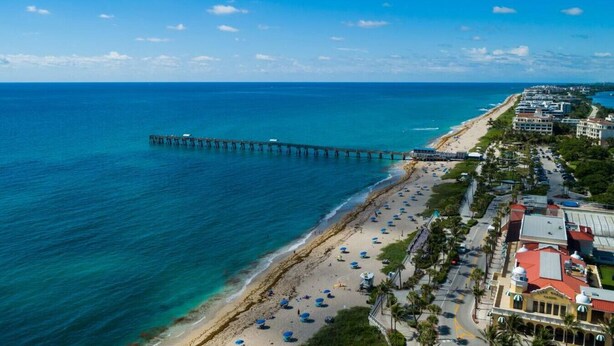 A coastal area with a long pier extending into the sea, a beach with umbrellas, and buildings along the shore.